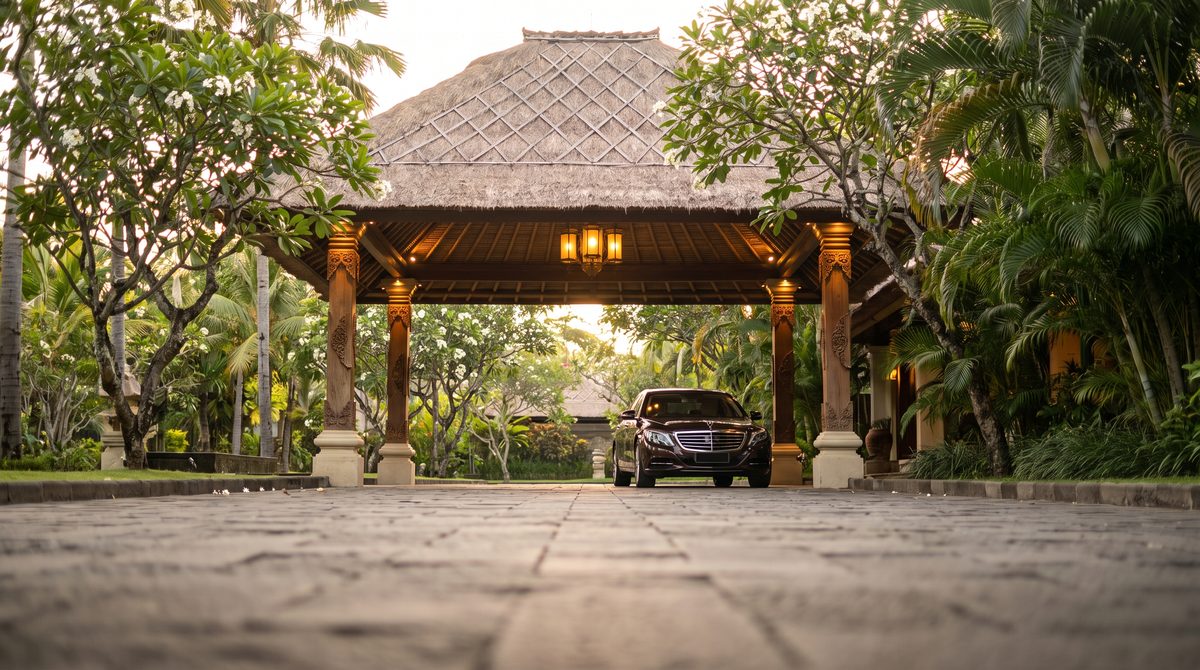 Thatched open-air arrival pavilion at a Balinese hotel at golden hour with carved timber columns, a lantern and a dark private transfer car waiting on stone paving among frangipani trees