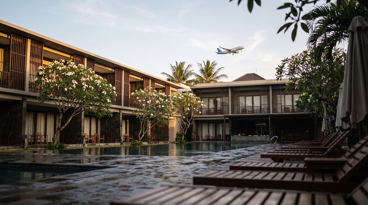 Low-rise Bali resort pool courtyard near the airport at early morning, with frangipani trees, dark timber screens and timber loungers beside a still pool, an airliner climbing in the pale sky above the palms