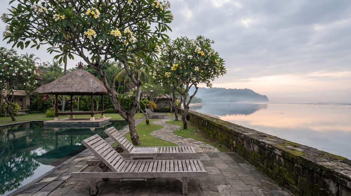 Quiet low-rise beachfront hotel garden in Sanur, Bali at soft early morning light with a thatched pavilion, timber loungers under frangipani trees and a still calm lagoon beyond