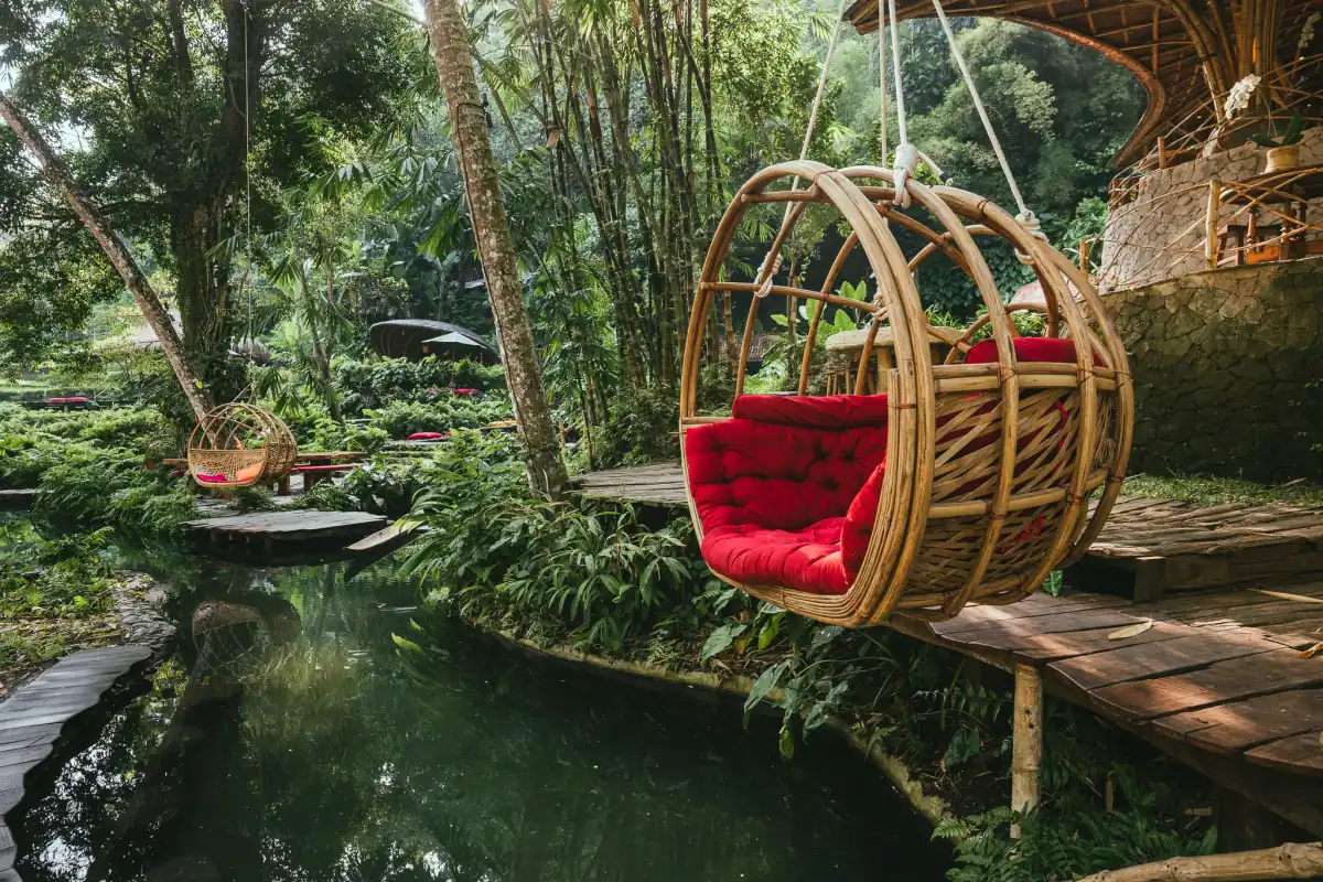 A rattan hanging seat with a red cushion beside a natural pool in the jungle at Bambu Indah